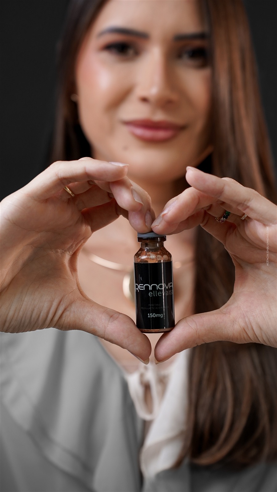 Fotografia de produto de estética: frasco em destaque entre as mãos formando um coração, rosto desfocado ao fundo